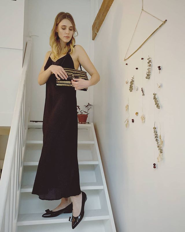 Gaia Weiss in a black sleeveless gown and black slippers carrying a side bag.